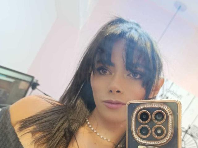 AnnaMooree, con capelli castano scuro, frangia e eyeliner, indossa una collana di perle, guarda la fotocamera, tiene in mano un telefono con una cornice dorata attorno alla fotocamera, labbra rosa..