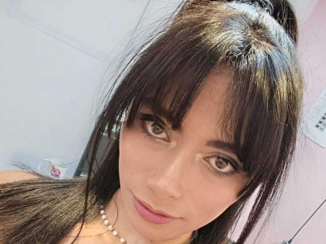 AnnaMooree a les cheveux brun foncé, avec une frange, et les yeux marron. Elle porte un collier de perles et du rouge à lèvres rose, avec un sourire discret. Ses seins sont partiellement visibles..