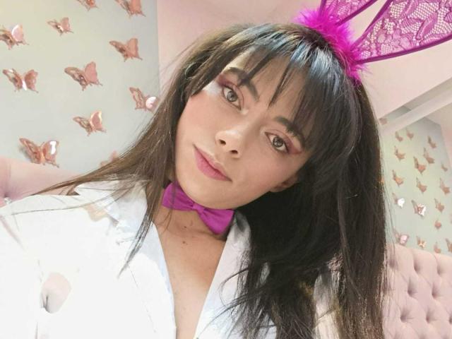AnnaMooree mit langem, schwarzem Haar, rosa Hasenohren und einem weißen Hemd, einem rosa Fliege, trägt Make-up, lächelt, schaut in die Kamera, zeigt ihre Brüste und posiert in einer süßen, verspielten Pose..