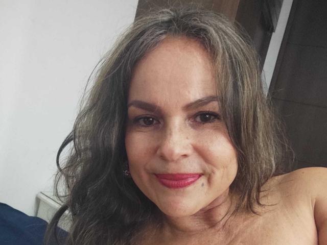 Danny69s tem cabelo grisalho e ondulado, olhos castanhos e batom vermelho. Ela está sorrindo com um leve franzido, mostrando seus seios de tamanho médio. Ela não está usando camisa, e o osso do colo está visível..