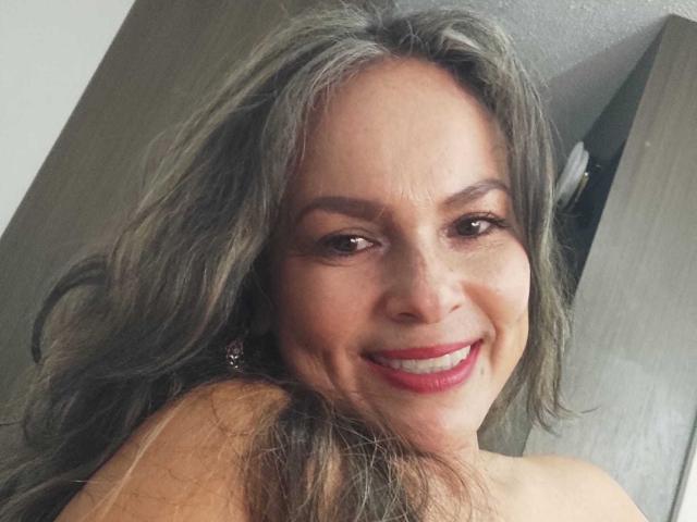 Danny69s tem cabelos grisalhos longos e ondulados, olhos castanhos e um sorriso que mostra batom vermelho, usa brincos visíveis e está sem camisa, exibindo os ombros nus..
