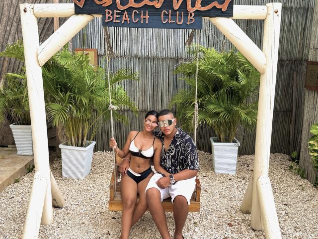MichelleXDominic y MichelleXDominic sentados en un columpio de madera, ella lleva un bikini blanco y negro, él lleva una camiseta negra y pantalones cortos blancos, ambos con gafas de sol..