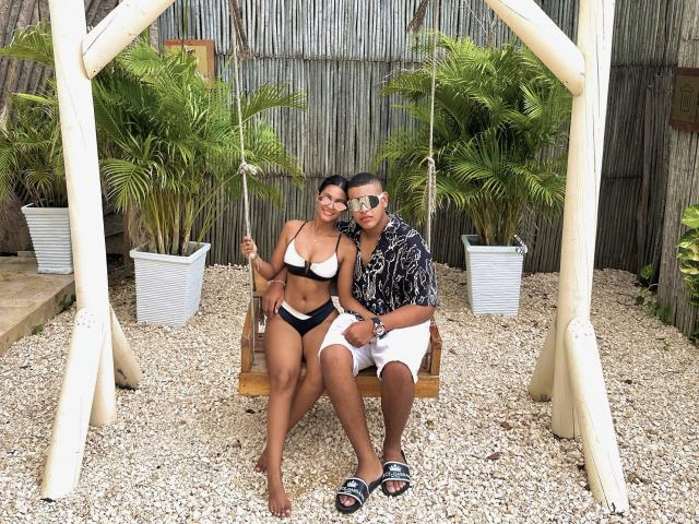 MichelleXDominic in einem schwarzen Bikini mit großen Brüsten, sitzt auf einer hölzernen Schaukel neben einem Mann in einem schwarzen Hemd, weißen Shorts und Sonnenbrille, auf Kies mit Palmen..