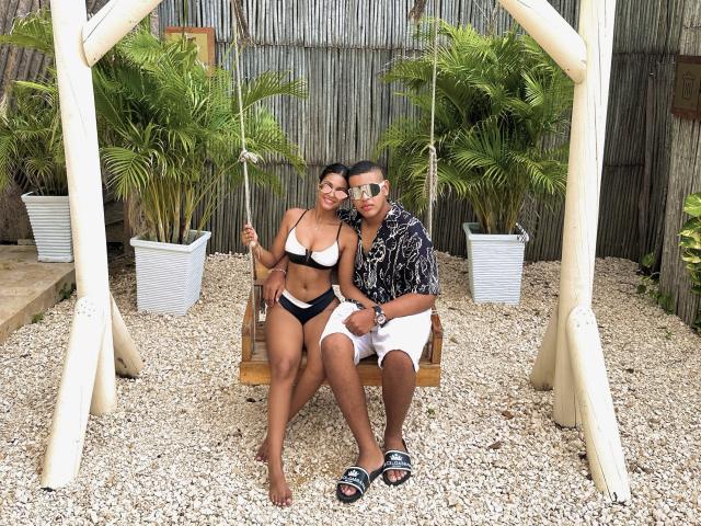 MichelleXDominic with tanned skin and dark hair, wearing a black and white bikini, sits on a swing with MichelleXDominic in a patterned shirt and white shorts, both smiling.
