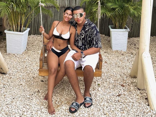 MichelleXDominic, avec une peau bronzée, un bikini noir et blanc, et de petites seins, assise sur une balançoire, avec MichelleXDominic en chemise noire, short blanc et sandales noires, souriant..
