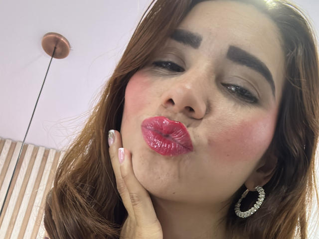 Bellasweeth's face, puffy pink lips, slightly upturned nose, brown eyebrows, pink blush, wearing a silver hoop earring, hand under chin, brown hair, pink nail polish, looking at camera.