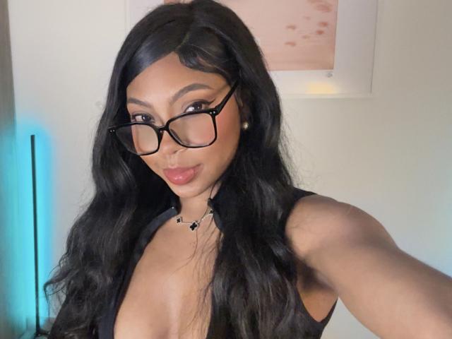 MissMaary with long black hair, wearing glasses, a deep V-neck black top showing her tits, and a star necklace, smiling at the camera.