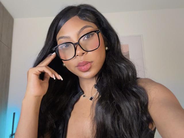 MissMaary with long black hair, wearing glasses, touching her cheek, pouty pink lips, and a necklace, showing her cleavage. She's looking at the camera with a slight smile.