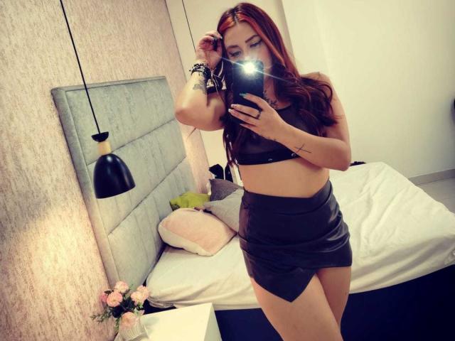 Camile Rouse mit roten Haaren, die ein schwarzes Leder-Crop-Top und einen passenden Rock trägt, macht ein Selfie vor einem Spiegel, wobei der Blitz ihrer Kamera blendet und ihre Hand ihre Haare berührt..