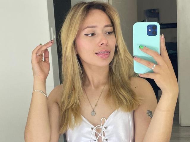 LinaMillia with blonde hair, white lace-up top, holding blue phone, tongue out, green nail, tattoo on right arm, silver bracelet, and necklace, taking mirror selfie.