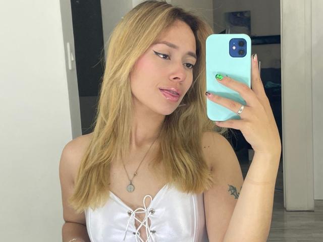 LinaMillia with blonde hair, white lace-up top, holding a blue phone, showing green nail, small tattoo on right arm, pink lips, ring on left hand, mirror selfie.