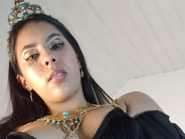 SoffiRousee with dark hair, wearing a jeweled crown, black off-shoulder top, and gold necklace with turquoise, looking at camera with winged eyeliner, earrings, and slightly parted lips.