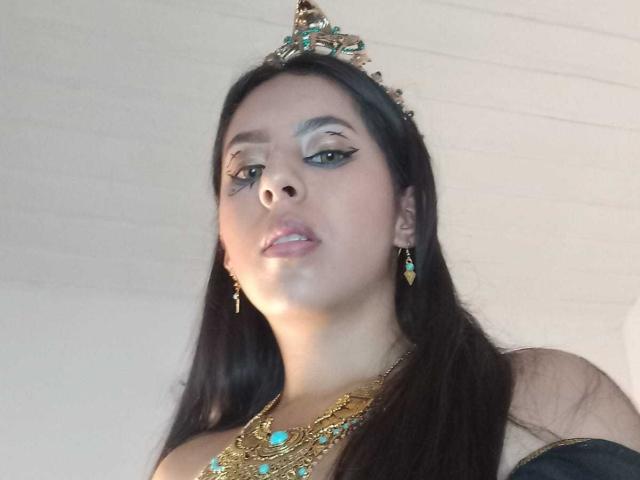 SoffiRousee with dark hair, green eyeliner, gold crown, teal earrings, and gold necklace with teal gems, looking down, lips slightly parted.