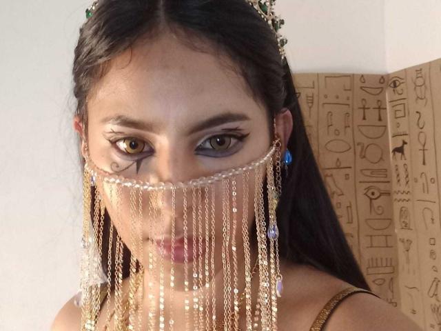 SoffiRousee with dark hair, wearing a gold chain face mask, green earrings, and a gold top, has brown eyes and a serious look. Beads hang down from the mask.