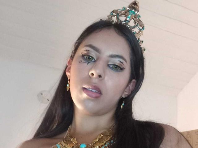 SoffiRousee has long black hair and wears a metallic tiara with turquoise and gold accents. She has green eyes, dark eyebrows, and light makeup with teal eyeliner. She wears gold and turquoise jewelry, including earrings and a necklace. Her expression is neutral, and she looks slightly downward. The image has a soft, slightly blurred effect. The lighting is even, highlighting her facial features and jewelry. The composition is a close-up, focusing on her face and upper shoulders.