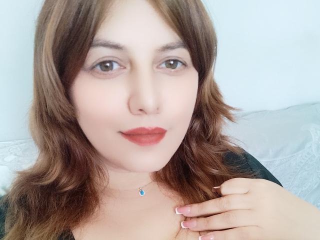 LoreyJolie has wavy brown hair, fair skin, brown eyes, red lipstick, and a black top. She's holding her chest with a hand, wearing a blue gem necklace.