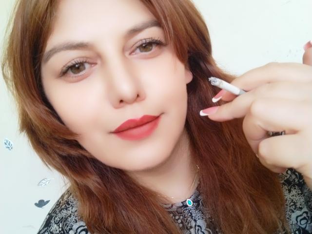 LoreyJolie with red lipstick, holding a cigarette to her right ear, has brown hair, light skin, and is wearing a patterned shirt, showing a slight smile.