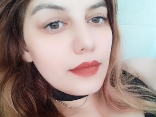 LoreyJolie has pale skin, brown eyes, red lipstick, and long brown hair with blonde highlights. She's wearing a black choker and a thin chain necklace.