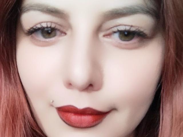 LoreyJolie's face with red lipstick, slight smile, green eyes, and a silver lip piercing on her lower left lip. She has pink hair and light skin.