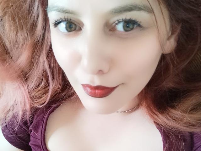 LoreyJolie with pale skin, red lipstick, and brown eyes, looking up at the camera. She has pinkish-brown hair, a purple top, and a thin necklace.