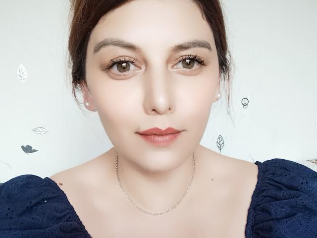 LoreyJolie's face, pale skin, brown eyes, slight nose piercing, subtle lipstick, dark blue textured top, pearl earrings, simple silver necklace, brown hair in a messy updo.