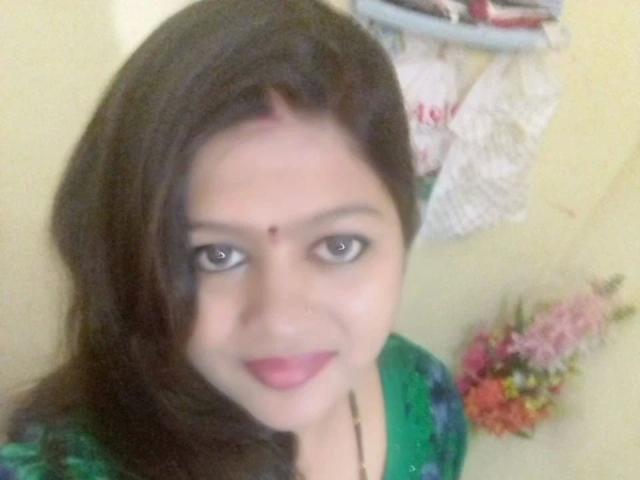 Nehaaahire-hot with dark hair, green top, nose ring, and red dot on forehead, smiling at camera. Boob-less, ass-free, pussy-free, soft-looking skin. Floral arrangement in background.