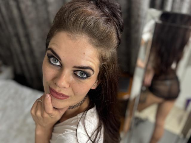 ChadeOnly69 with dark brown hair in a messy bun, blue eyes, and black eyeliner, wearing a white top, holding her chin, looking at a mirror reflecting her ass.
