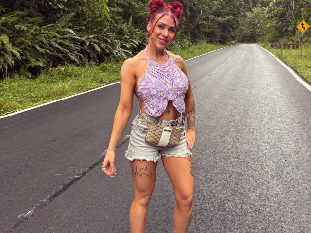 A photograph of JustSophia-hot standing on a wet, empty road. She has a medium build with tattoos on her right thigh. Her hair is dyed red and styled in two space buns. She's wearing a purple, crocheted, halter-top that reveals her midriff and high-waisted denim shorts with a quilted belt. Her tattoo on the right thigh is a simple outline. The road is bordered by lush greenery. She's smiling and looking directly at the camera. The wet road reflects light, giving it a shiny texture.