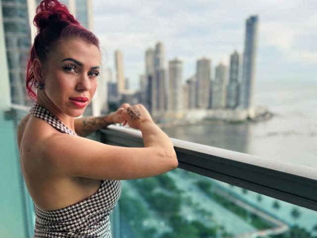 JustSophia-hot stands on a balcony with her arms resting on the railing. She has bright red hair in a bun, heavy eyeliner, and red lipstick. She wears a black-and-white houndstooth-patterned sleeveless dress. Her tattoos are visible on her arms. The cityscape in the background shows tall buildings near a waterfront. The sky is cloudy, and the balcony overlooks green spaces and a river. Her expression is confident, and she looks directly at the camera. The image is sharply focused on her with a slightly blurred background.