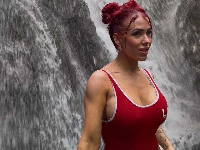JustSophia – attraktiv mit roten Haaren zu Dutt, trägt ein rotes Sport-Tanktop, zeigt ihre Brüste, steht vor einem Wasserfall und schaut in die Kamera..