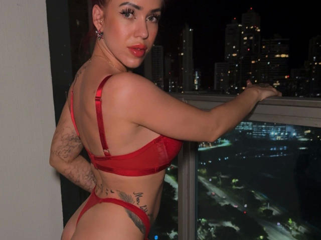 JustSophia - sensual, vestindo lingerie vermelha, mostrando os seios e o bumbum, tatuagens visíveis, batom vermelho, luzes da cidade ao fundo, em pé perto de uma varanda à noite, olhando para trás..