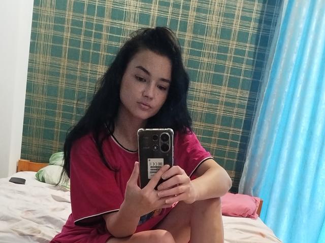 TishaUrsa, con cabello negro largo, pecas y una camiseta rosa, sentada en la cama, sosteniendo un teléfono, tomando una selfie en el espejo, una cortina azul a la derecha, una pared verde con estampado detrás..