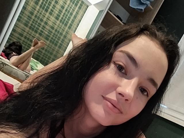 TishaUrsa, com cabelos pretos longos, pele clara e maquiagem discreta, está deitada em uma cama, com seus pequenos seios visíveis, olhando para a câmera com um leve sorriso. Suas pernas estão levantadas..