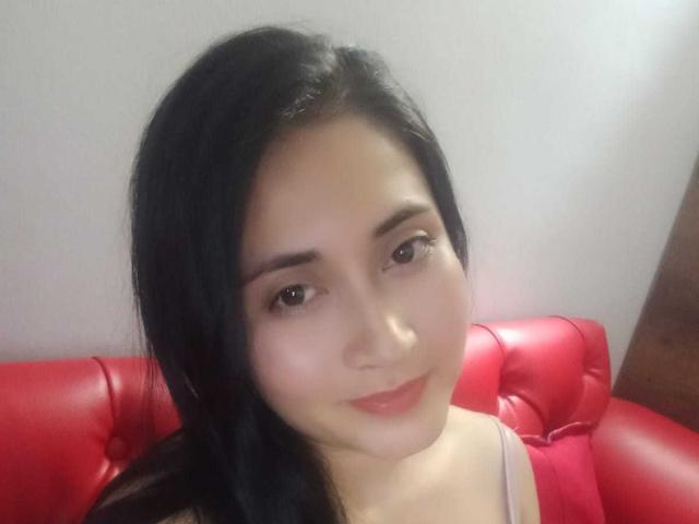 CatalinaGrey has black hair, light skin, and a slight smile. She's sitting on a red leather couch, wearing a grey top, with her tits partially visible.