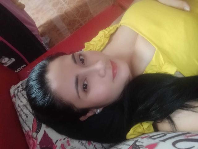 CatalinaGrey with black hair, yellow top, and pink lipstick, lying on a red bed, smiling at the camera, showing her small tits and cute face.