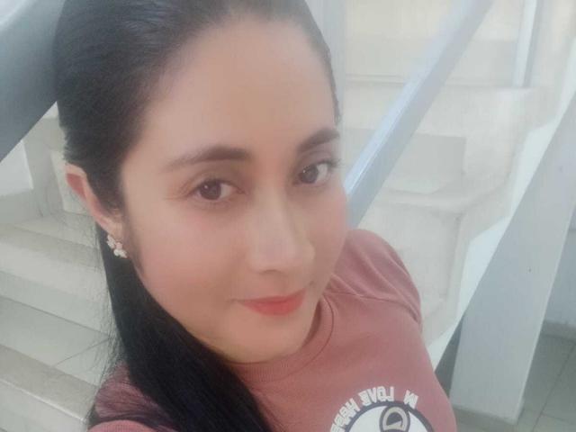 CatalinaGrey has light brown skin, dark brown eyes, and long black hair. She's wearing a pink shirt, smiling, and has small earrings. Her tits are slightly visible.