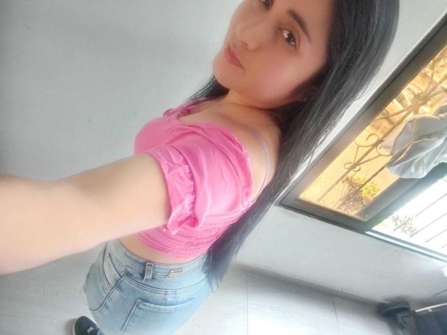 CatalinaGrey with long black hair, pink off-shoulder top, and blue jeans, showing her small tits and round ass in a selfie. She's looking at the camera, slightly smiling.