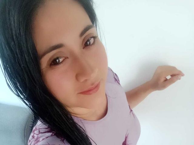 CatalinaGrey has black hair, brown eyes, and a light skin tone. She's wearing a purple top, looking up with a slight smile, and her right arm is raised.