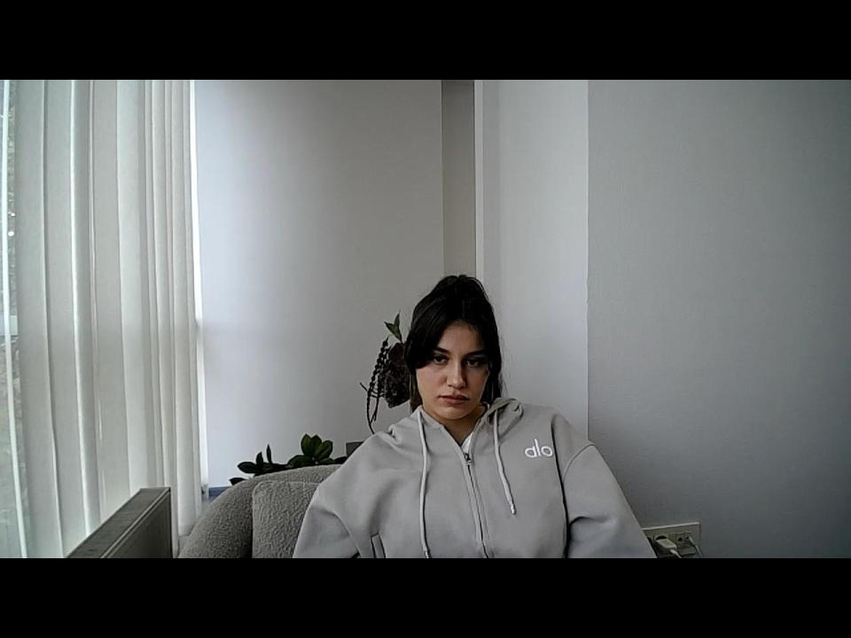 Flaaina sits in a grey chair against a white wall, wearing a light grey Adidas hoodie with a small logo on the left chest. She has dark, wavy hair and a neutral expression. Light from a window with vertical blinds casts soft shadows. A small green plant is on the chair's left. The image is a photograph with a subtle, realistic style.