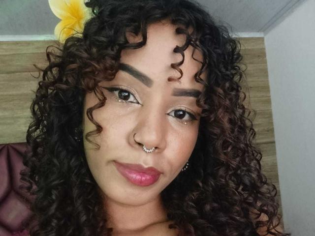 MiissDiiamondss has curly brown hair, dark eyebrows, a nose ring, and pink lipstick. She's looking at the camera with a slight smile. Her skin is light brown.