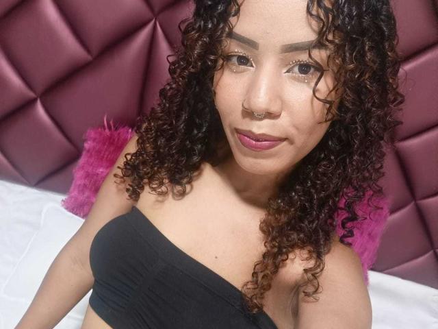 MiissDiiamondss with curly brown hair, nose ring, and pink lipstick, wearing a black strapless top, sitting on a bed with pink pillows, smiling at the camera.