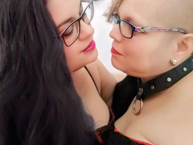 Two LatinoHardGirls with glasses, one with long black hair and red lipstick, the other with blonde hair and black collar, gently touching faces, showing cleavage and soft expressions.