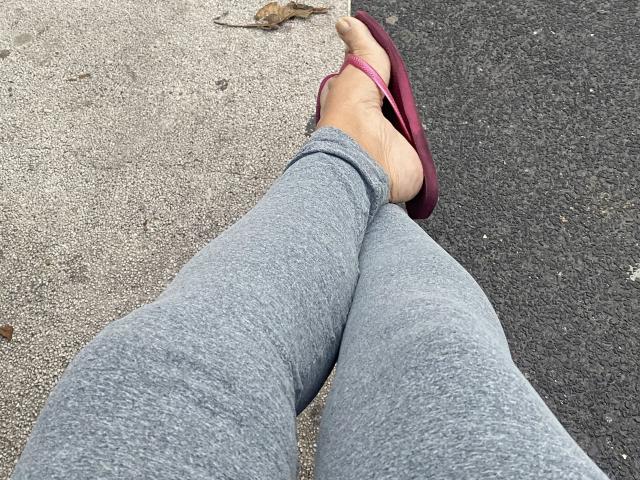 Brunha's legs in gray pants with pink flip-flops on asphalt, showing bare feet with a small rock near the top. Her toes are slightly curled.
