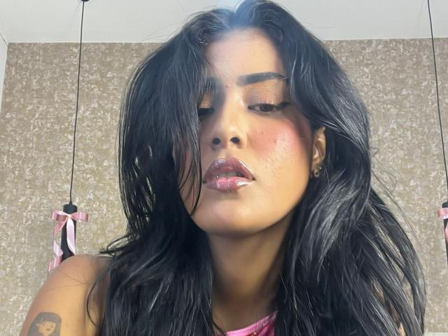 Babilett, con labios rosados y brillantes, cabello oscuro y húmedo, y un tatuaje en su brazo derecho, vestida con una blusa rosa, tiene el rostro sonrojado y los labios ligeramente entreabiertos y brillantes..
