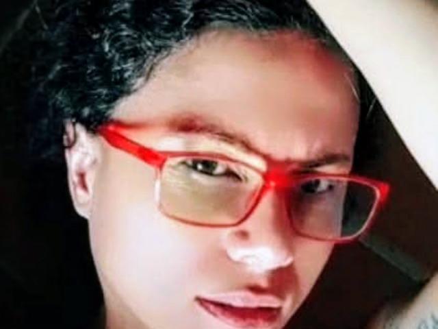 RosannaBlue with curly black hair, wearing bright red glasses, looking slightly to the side with a subtle smile, has light brown skin and green eyes.