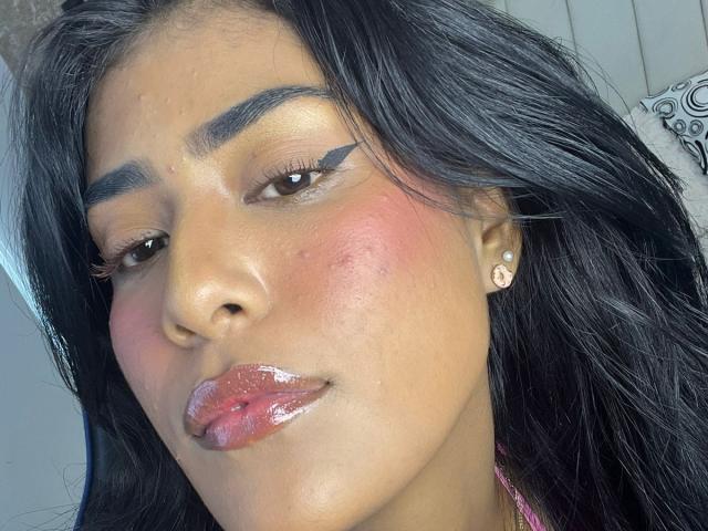 Babilett has shiny pink lips, a small zit on her right cheek, and glossy black hair. She's wearing diamond earrings and has thick black eyebrows with winged eyeliner.