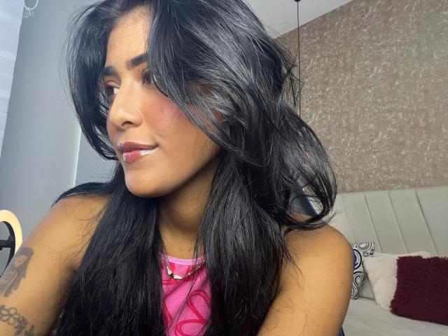 Babilett has long black hair, brown skin, and pink lipstick, wearing a pink top, looking to the side with a slight smile, showing a tattoo on her right arm.