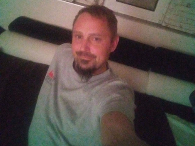 Major-hot with a short brown beard and mustache, wearing a gray t-shirt, smiling at the camera, sitting on a black and white bench.