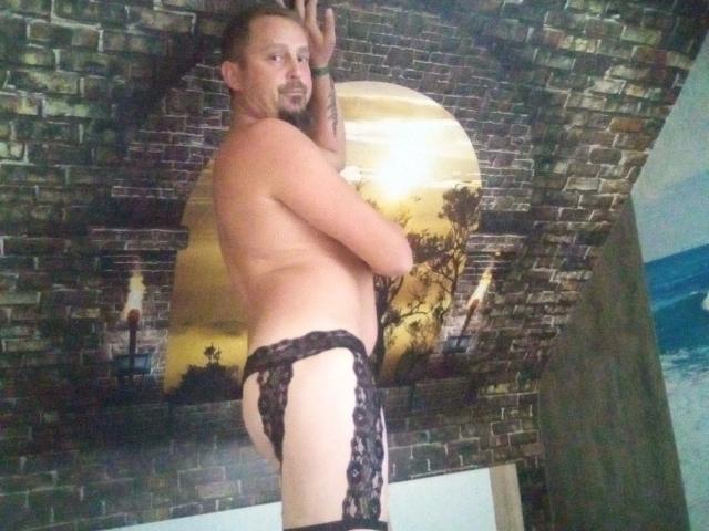 Major-hot, shirtless, with a lace thong, has a muscular butt, tattoo on left arm, and short brown hair, smiling at the camera. Ceiling has a brick pattern.