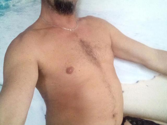 Major-hot's shirtless upper body, showing a hairy chest, small brown nipple, and a thin necklace. His arm is outstretched, and his face is partially visible with a beard.